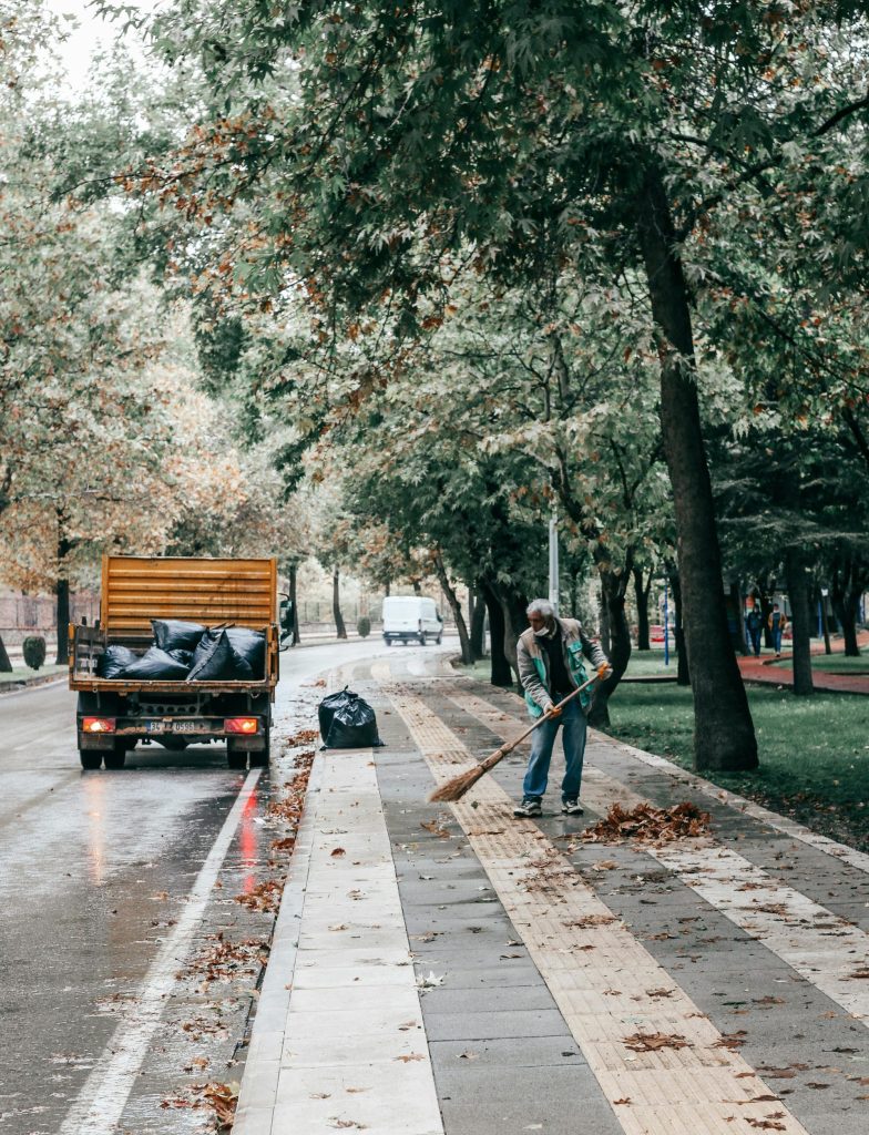 A man sweeping fallen leaves on a rainy tree-lined street with a parked truck nearby.